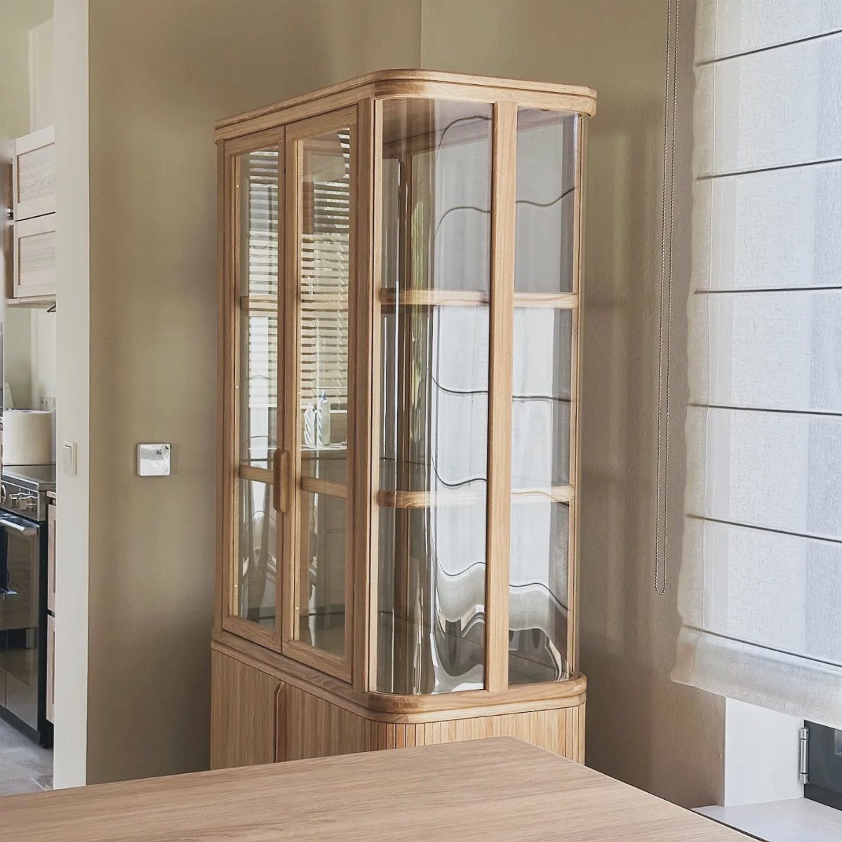 Light wood kitchen cabinet with curved edges and glass-front doors against beige walls.
