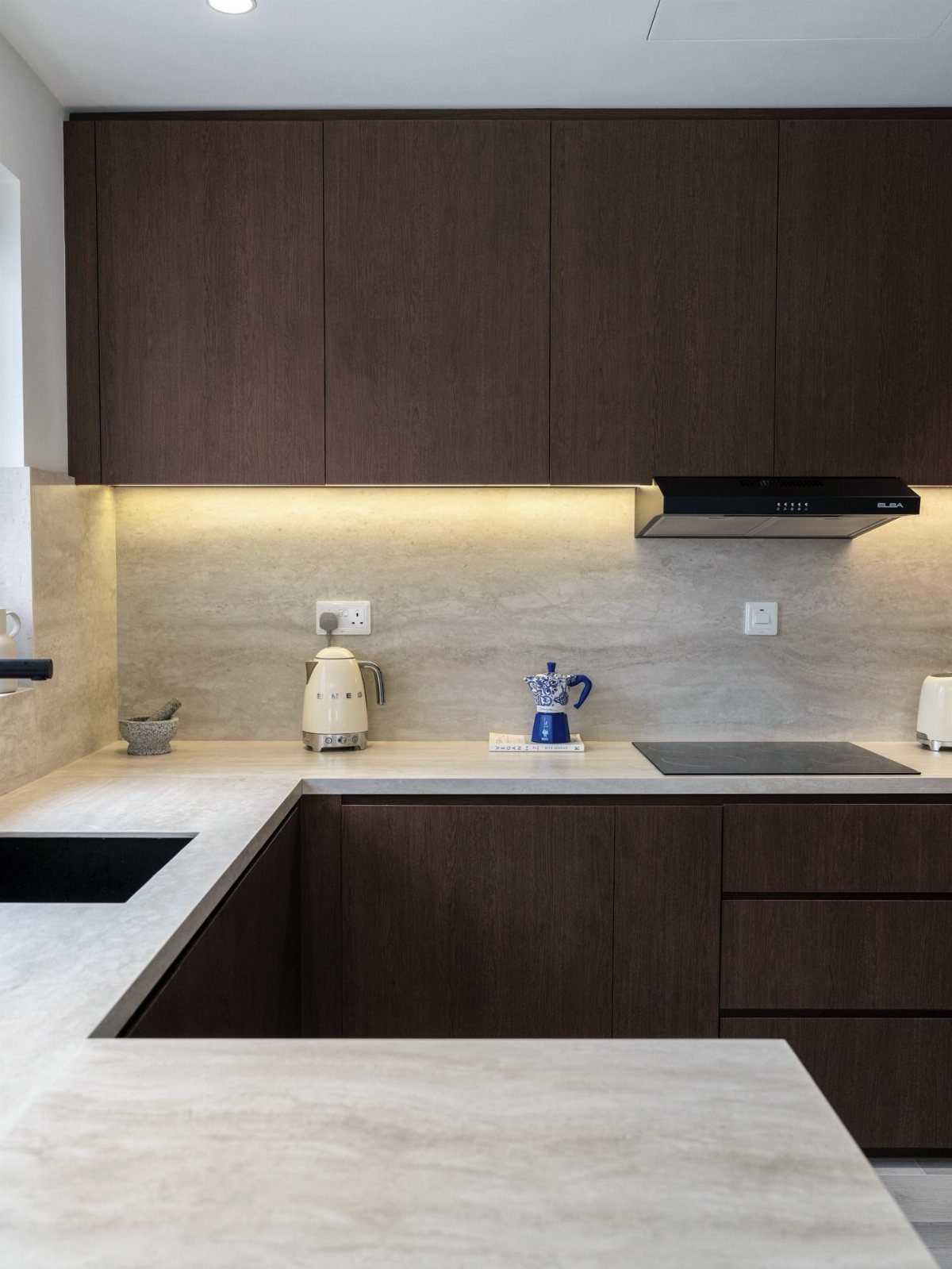Chocolate brown flat-panel kitchen cabinets with light beige stone countertops and under-cabinet lighting.