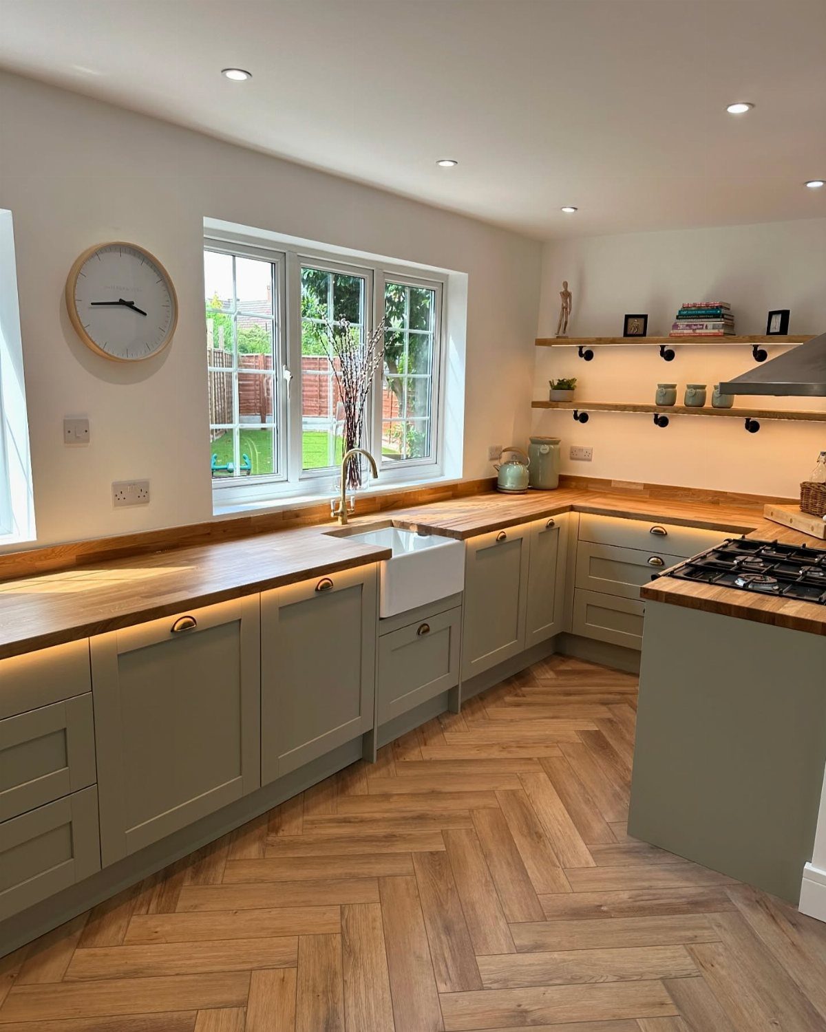Sage green kitchen cabinets with light wood countertops and herringbone wood flooring.