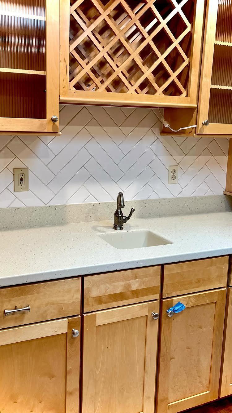 White herringbone tile backsplash behind a white kitchen counter and light wood cabinets.